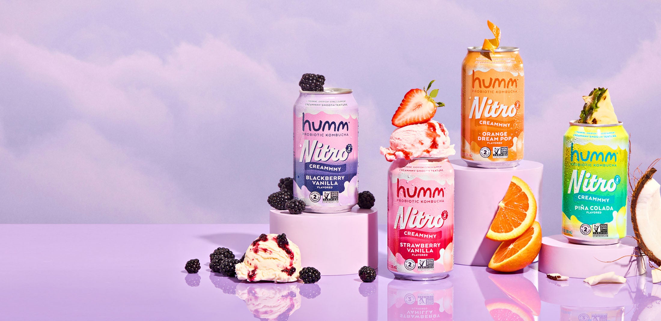 Four cans of Humm Nitro drinks in different flavors—Blackberry Vanilla, Strawberry Vanilla, Orange Creamsicle, and Piña Colada—are displayed on a pastel purple surface with matching cylindrical stands. Each can is garnished with corresponding fruits and ice cream scoops: blackberries and a scoop with berries for Blackberry Vanilla, strawberries and a scoop for Strawberry Vanilla, orange slices for Orange Creamsicle, and pineapple pieces with coconut for Piña Colada.