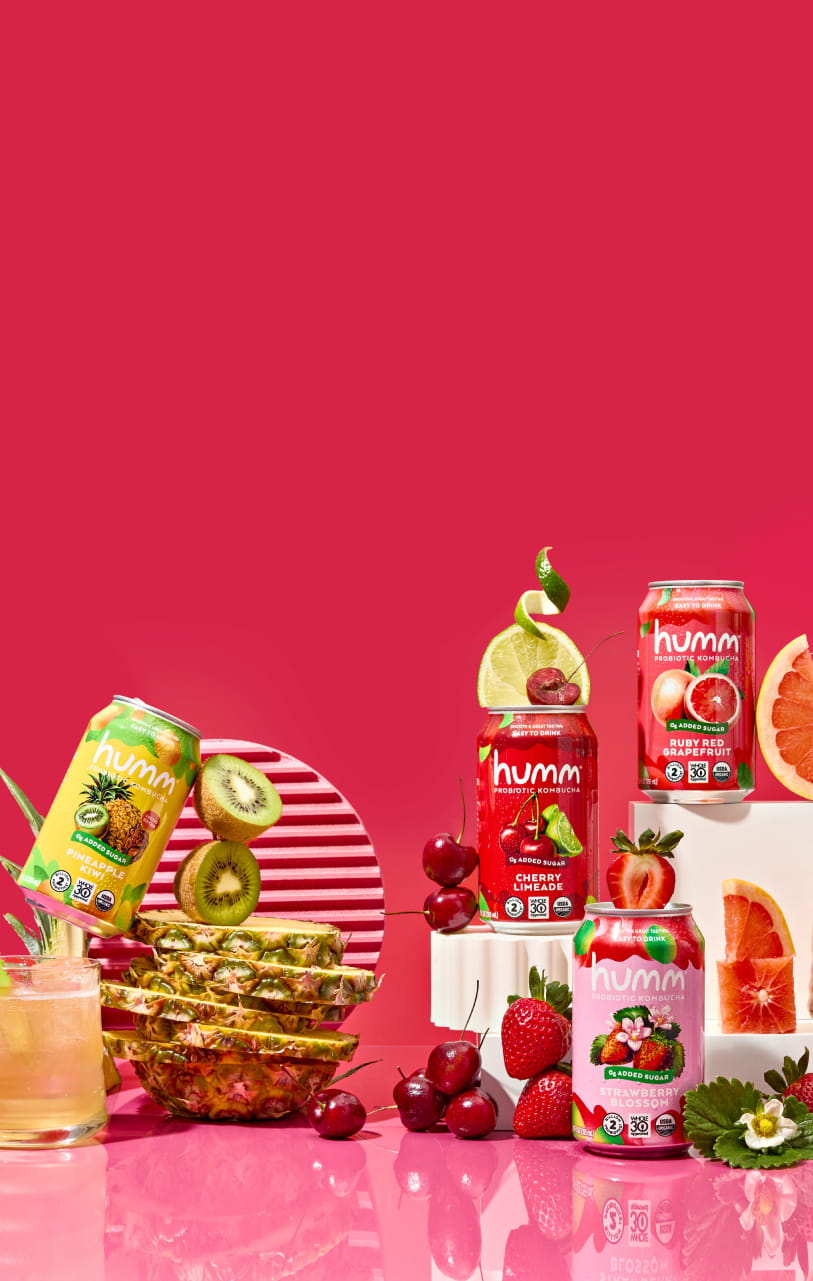 A vibrant display of Humm Probiotic Kombucha cans in various flavors on a pink surface with a red background. From left to right: a glass with kombucha and aloe garnish, a Pineapple Kiwi can with kiwi slices and pineapple chunks, a Cherry Limeade can with cherries and lime slices, a Ruby Red Grapefruit can with grapefruit wedges, and a Strawberry Blossom can with strawberries and blossoms. The cans are arranged on white blocks with fruit accents.